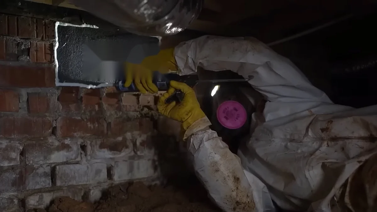 Technician in a protective suit and respirator sealing insulation board on a brick crawl space wall